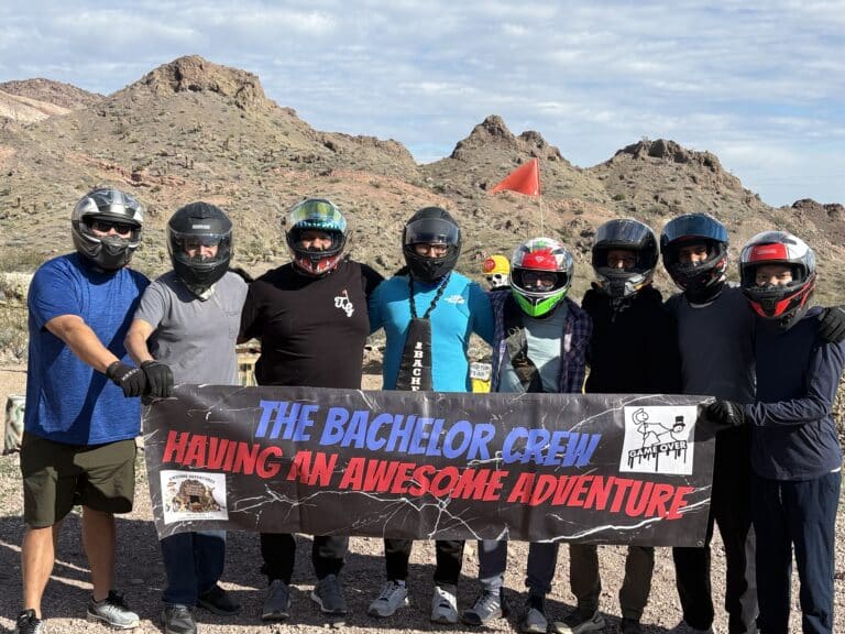 Group in helmets and outdoor gear by Nevada desert hills, banner reads: THE BACHELOR CREW HAVING AN AWESOME ADVENTURE; Las Vegas ATV tour.