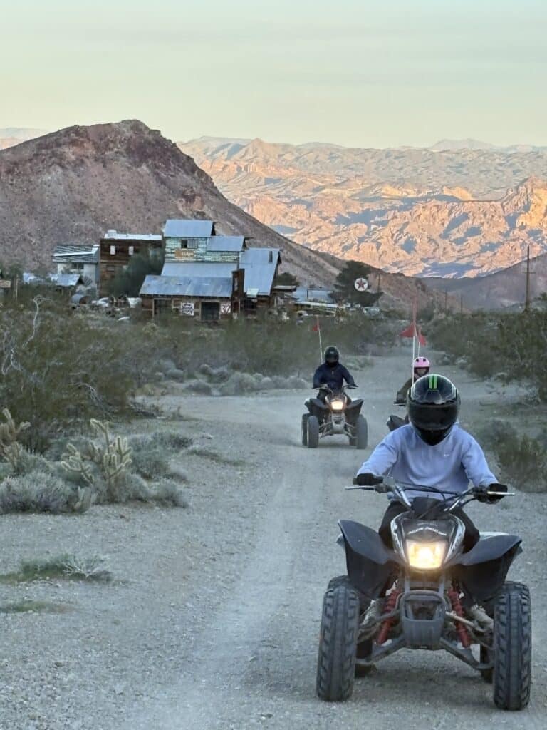 Helmeted riders drive ATVs toward the camera on a Nevada desert trail near rustic ghost towns and rocky mountains at sunset.