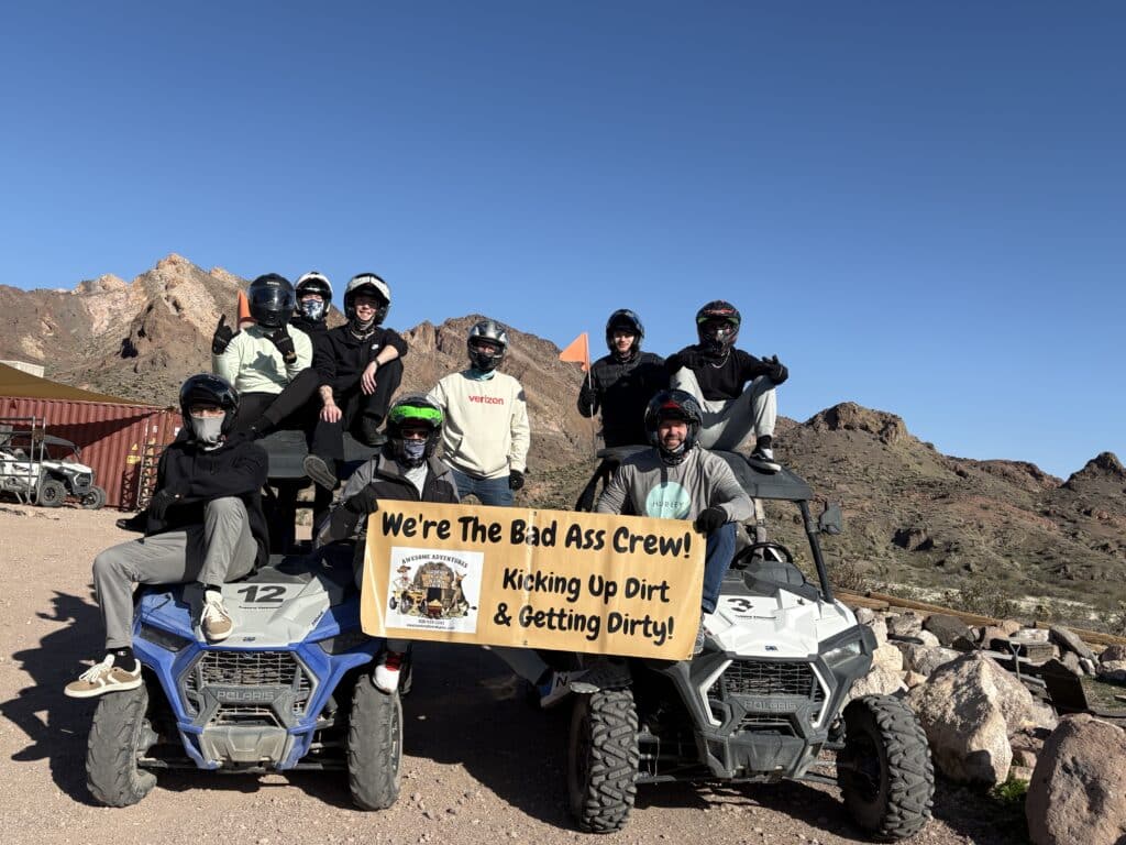 Adventure-seekers pose on ATVs in Nevada desert near Las Vegas, holding sign on RZR tour under blue sky.