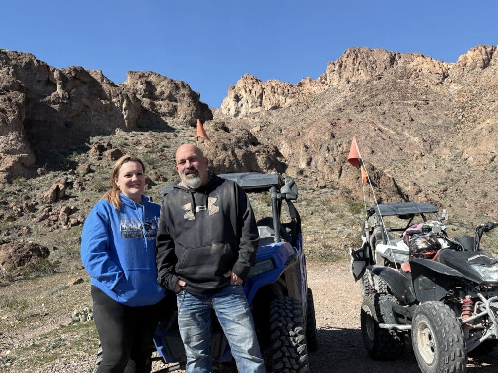 Couple on a Nevada ATV tour stands by off-road vehicles, rocky trail, and sunlit mountains near Las Vegas under clear blue sky.