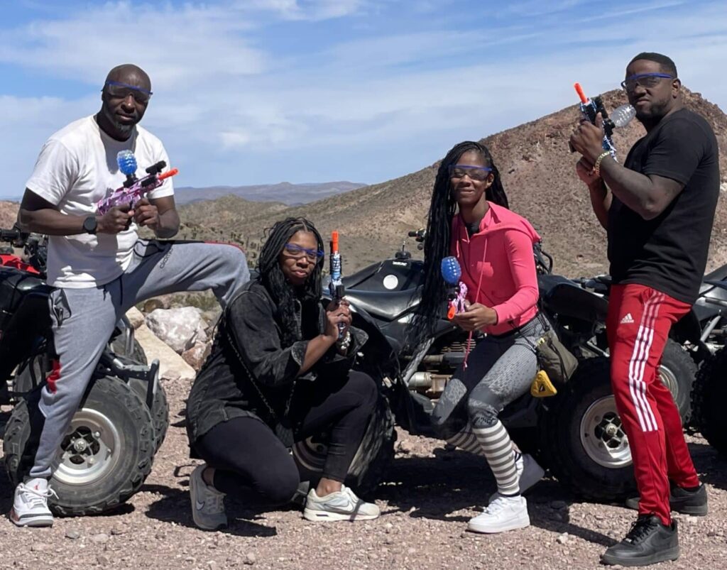 Group poses with toy guns & goggles by ATVs on a Colorado River RZR tour near Las Vegas, Nevada, with desert and mountain views.