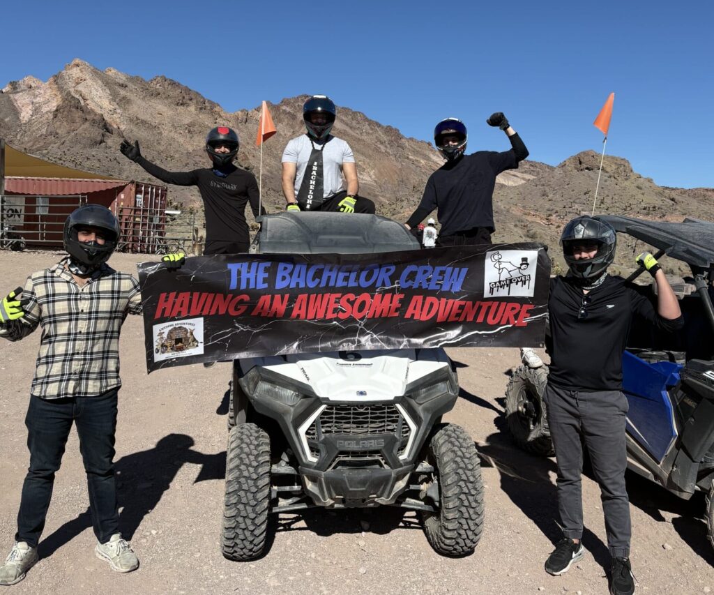 Group in helmets poses by an ATV in Nevada desert with a “Bachelor Crew” banner, ready for Las Vegas RZR tours near Colorado River.