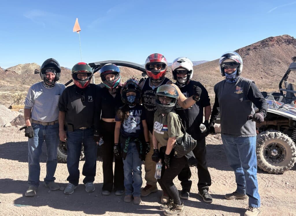 Eight adventurers in helmets pose by their RZR in Nevada desert near Las Vegas, set for an ATV tour and off-road adventure.