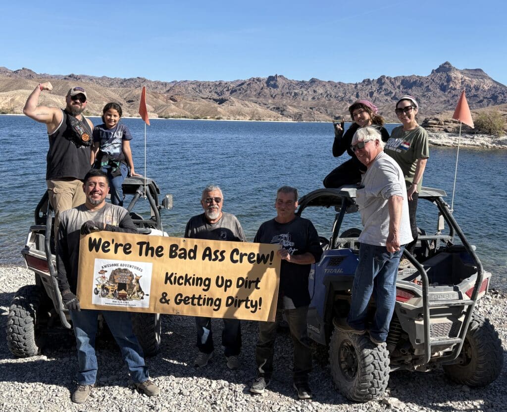 Nine people, two ATVs, and a “Bad Ass Crew” banner pose by a Nevada lake on a Ghost Town ATV tour near Las Vegas.