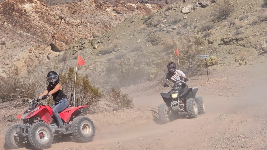 Helmeted riders on ATVs with red flags explore dusty Eldorado Canyon near Las Vegas, Nevada—ATV tours amid rocky desert hills.