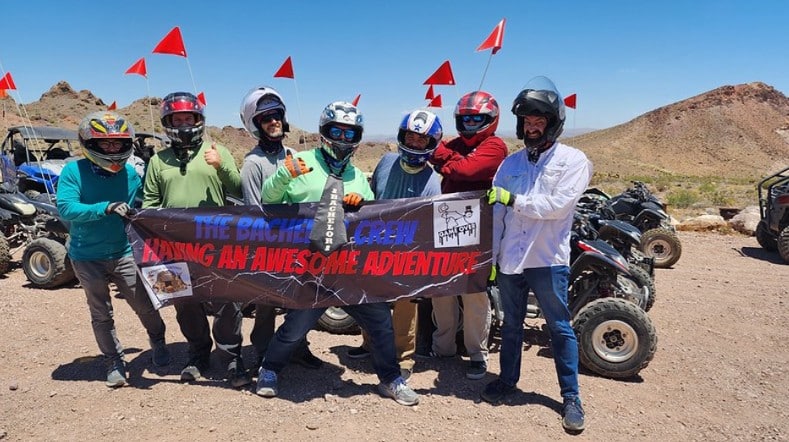 Seven friends with helmets and gear pose by ATVs in the Nevada desert on a Las Vegas ATV tour adventure near red rocky hills.