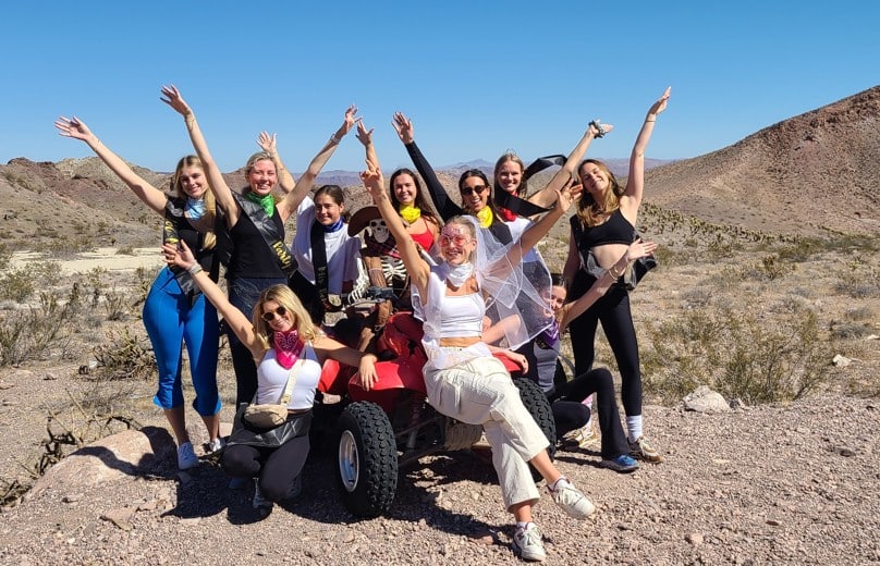 Smiling women on a Las Vegas RZR ATV tour pose by a red vehicle in Nevada desert, enjoying off-road adventure under blue skies.
