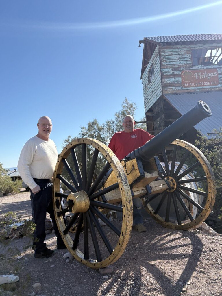 Two men by a black and gold cannon at a Nevada ghost town, near rustic wood buildings, perfect for Las Vegas sightseeing tours.