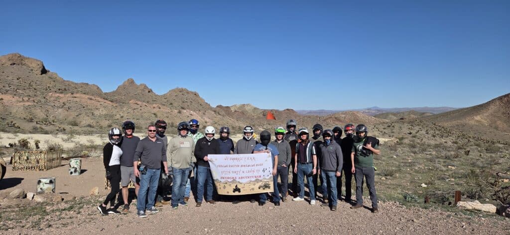 Adventure group in helmets holds sign on Nevada rocky terrain near Las Vegas, ready for RZR ATV tour by the Colorado River.