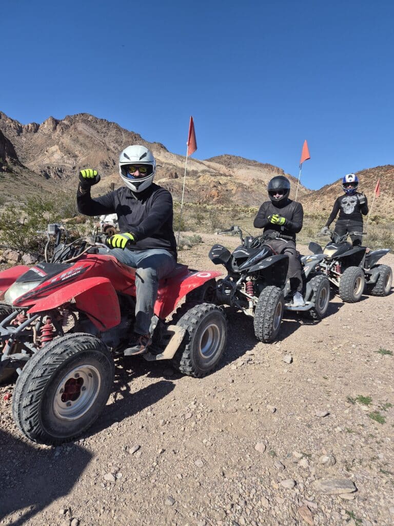 Adventurers on an ATV tour near the Colorado River in Nevada ride a rocky trail, red flags flying, lead rider raising a fist.