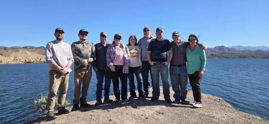 Nine friends pose on a rocky Nevada lakeshore near the Colorado River, mountains behind them, after an exciting ATV tour.