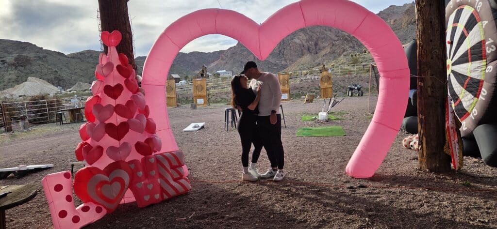 A couple kisses under a pink heart arch near the Colorado River, with Nevada mountains and a giant dartboard as ATV tours await.