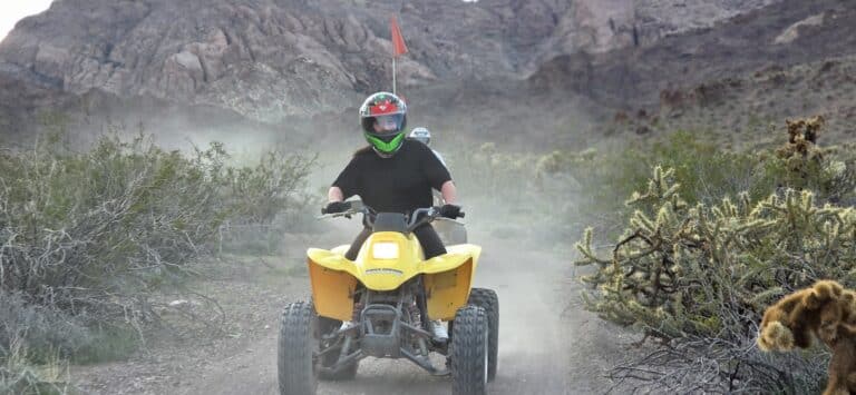 Helmeted rider on a yellow ATV explores a dusty Nevada desert trail near Las Vegas with cacti, mountains, and ATV tour adventures.