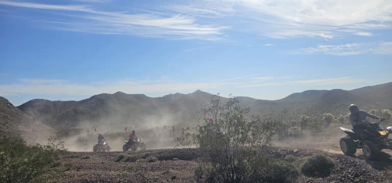 Adventurers ride ATVs on a dusty Nevada desert trail near Las Vegas, with mountains and shrubs under bright blue sky.