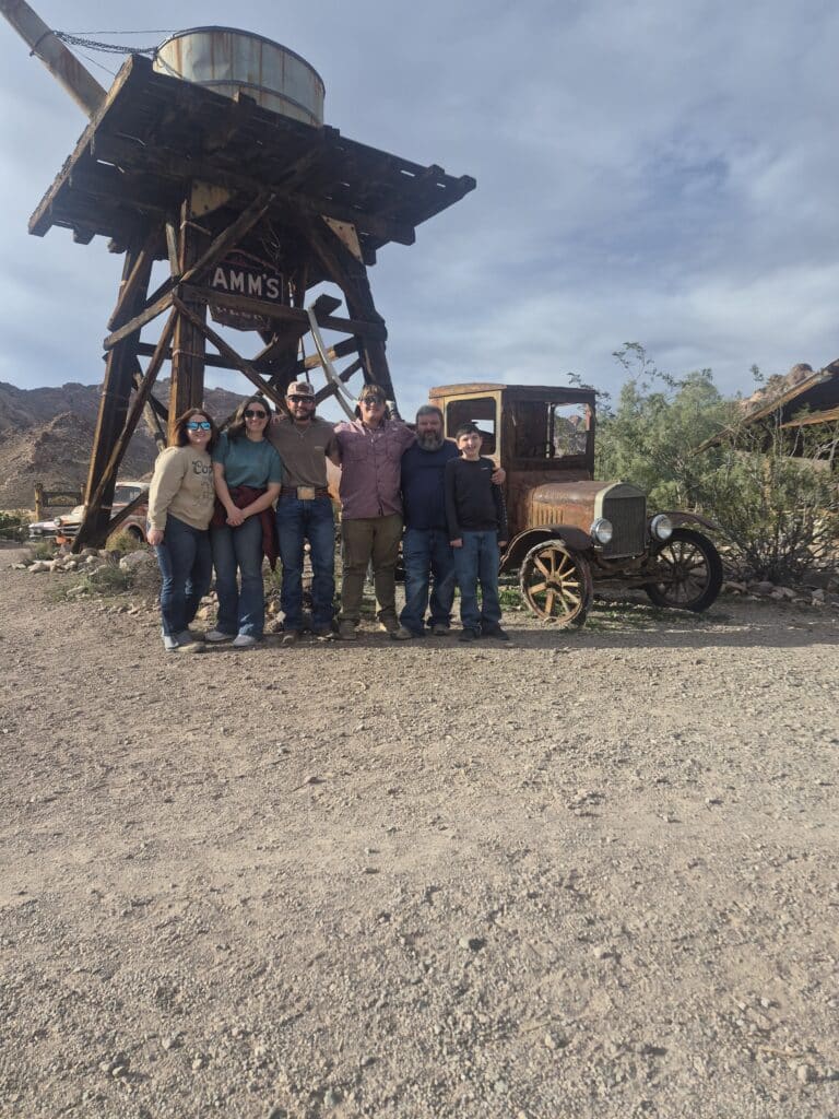 Group smiling by a wooden water tower, vintage car in a Nevada ghost town near the Colorado River—Las Vegas ATV tour fun.