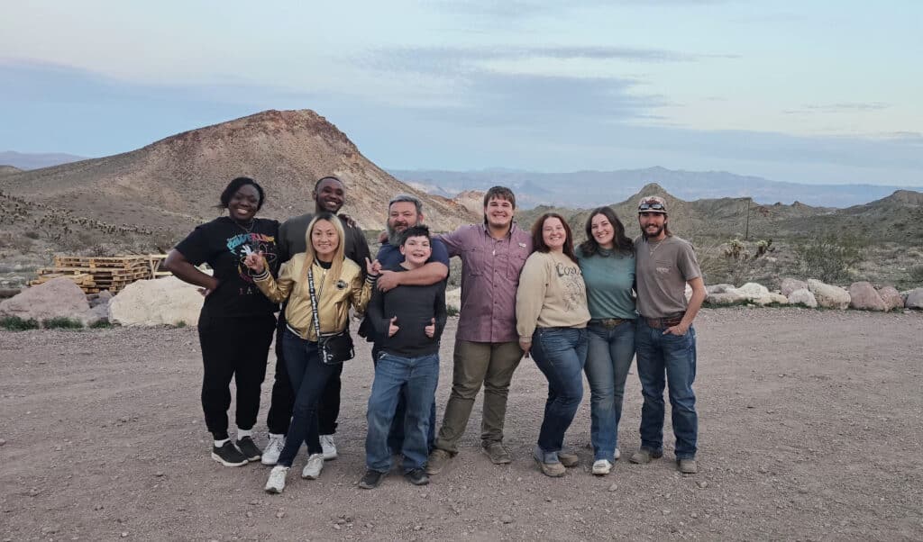 Eight friends smile on a Nevada dirt road at dusk, mountains behind them after an ATV tour near the Colorado River, Las Vegas.