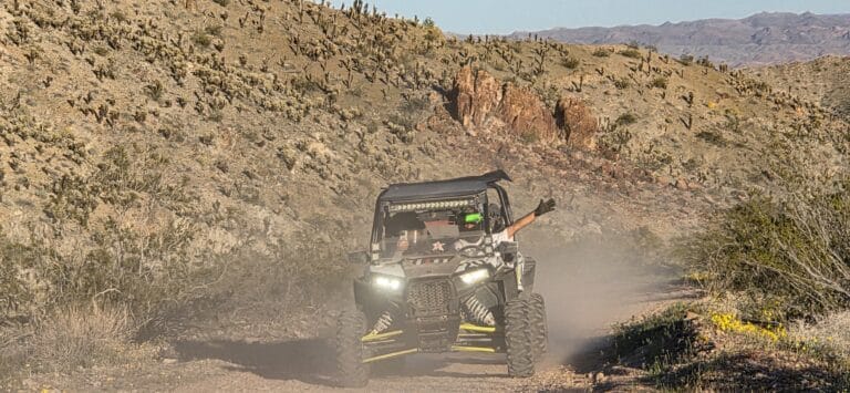 Person enjoying a RZR off-road ride near Las Vegas, Nevada, raising an arm while driving past cacti and rocky desert hills.