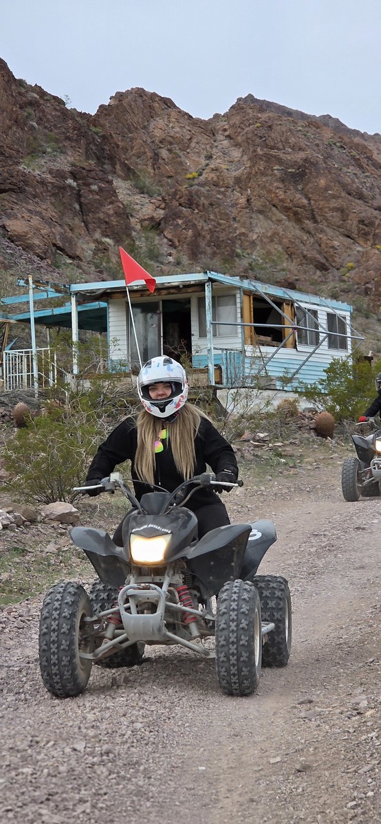 ATV rider with red flag explores Nevada dirt trail near rocky hills, blue house, and another ATV—perfect for Las Vegas Ghost Town tours.