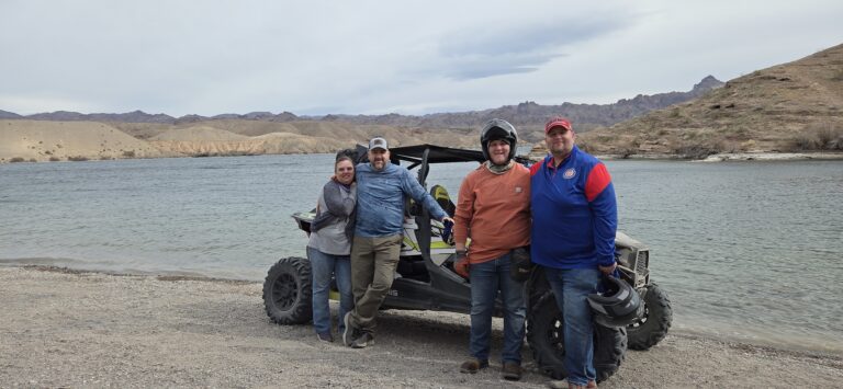 Four adults smile on a rocky Nevada beach by an ATV, with Colorado River and desert hills near Las Vegas in the background.