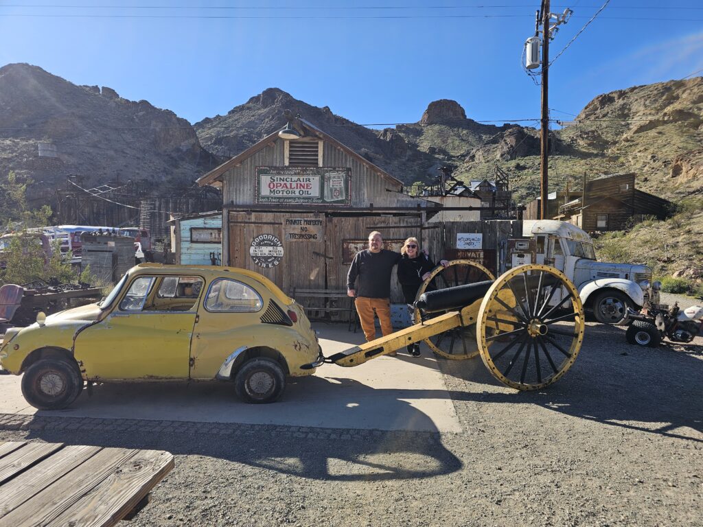 Tourists in Nevada’s Ghost Town pose by a quirky yellow car near vintage signs, old vehicles, and desert mountains near Las Vegas.