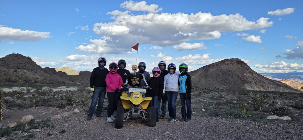 Eight helmeted riders by a yellow ATV on a Nevada desert tour near Las Vegas, with mountains and blue sky in the background.