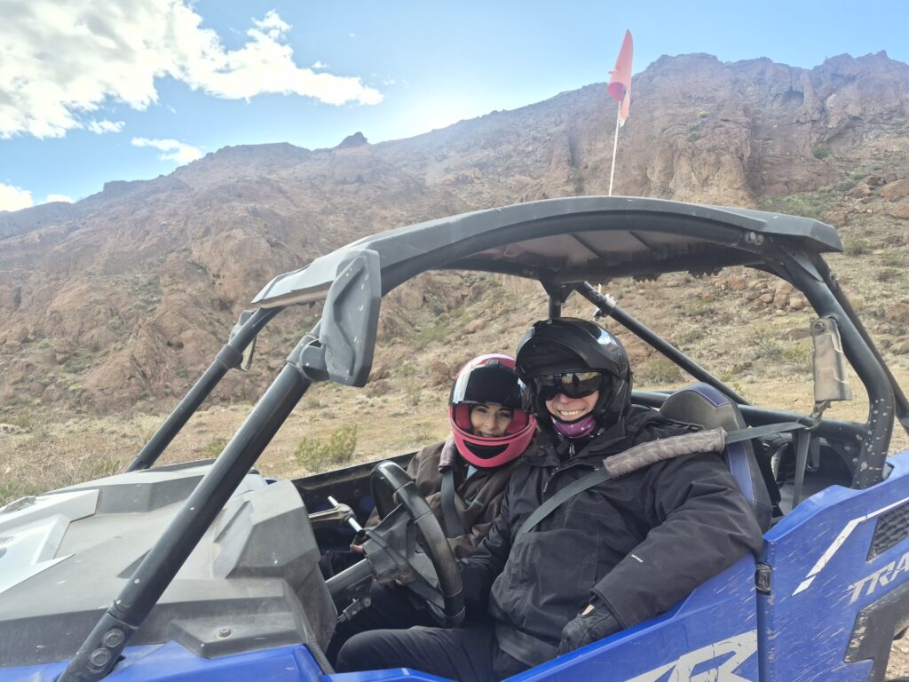 Smiling riders in helmets enjoy a blue RZR off-road ride near Las Vegas with Nevada mountains and clear skies in the background.