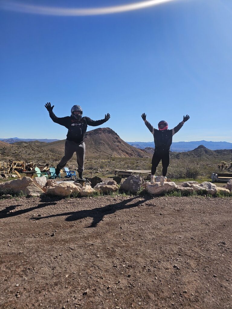 Adventurers in helmets jump with arms raised on a Nevada ATV trail near desert mountains under the clear blue sky.