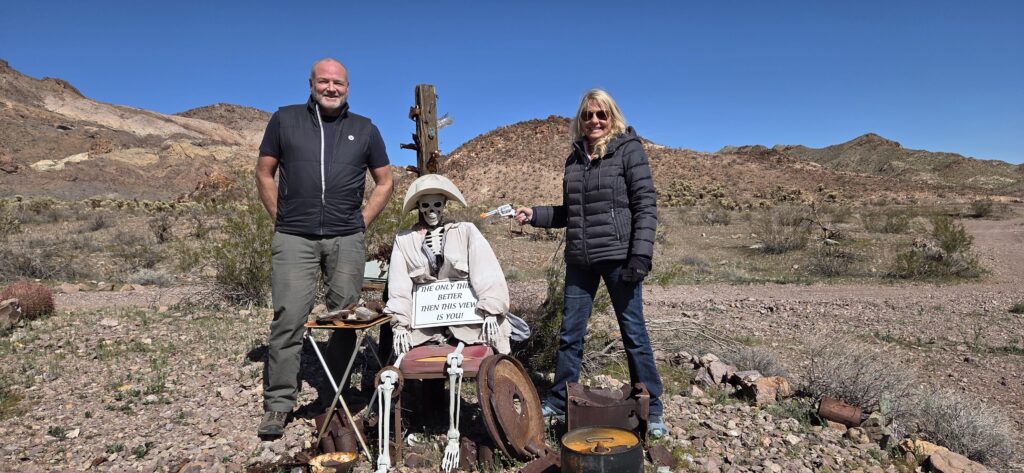Couple poses in Nevada desert by a skeleton in a hat near Ghost Town, mountains and sky behind; perfect for Las Vegas sightseeing tours.