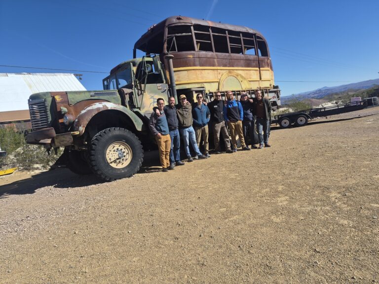 Group awaits ATV tour by a rusty double-decker in Nevada desert near mountains, trailers, and clear skies.