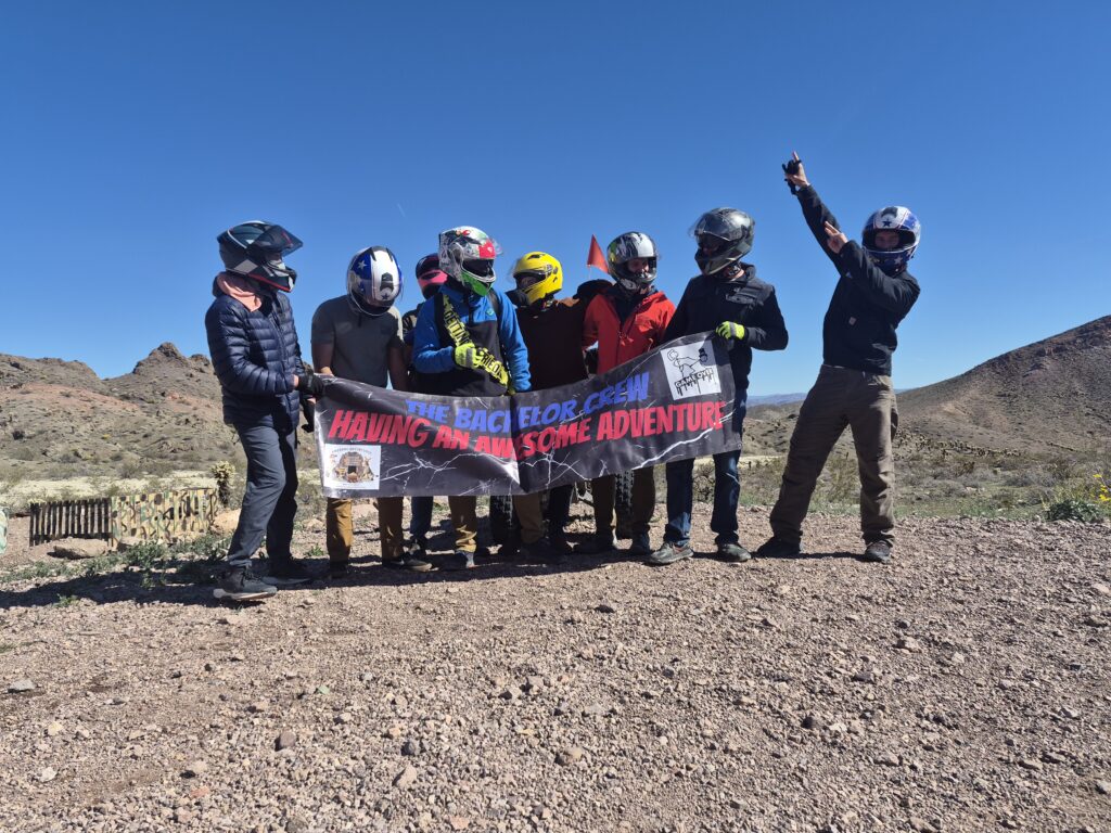 Adventurers in motorcycle gear pose by the Colorado River near Las Vegas, holding a banner; mountains and blue sky behind.