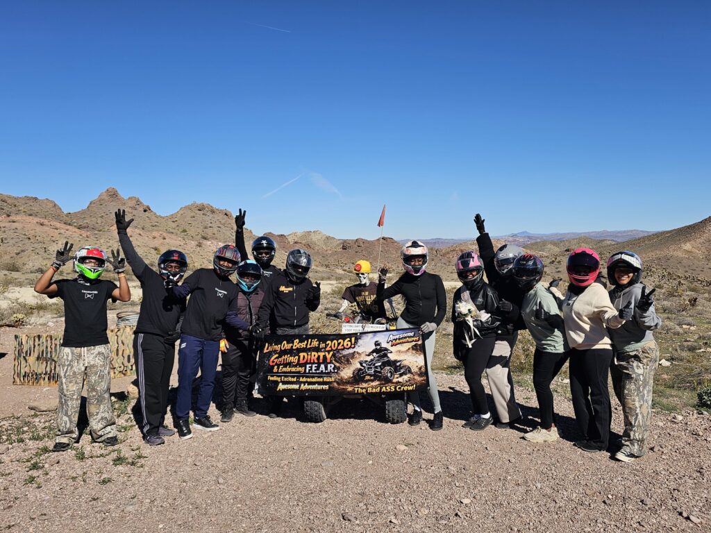 Adventurers in helmets raise hands on Nevada rocky terrain, holding a "Getting DIRTY, Kicking FEAR!" banner near Las Vegas.