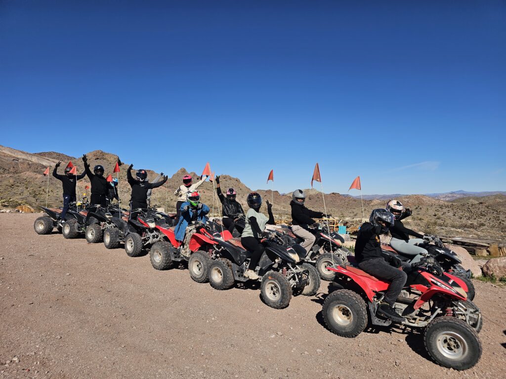 Riders in helmets line up on red ATVs with orange flags for a Nevada ATV tour near Las Vegas, mountains and blue sky behind.
