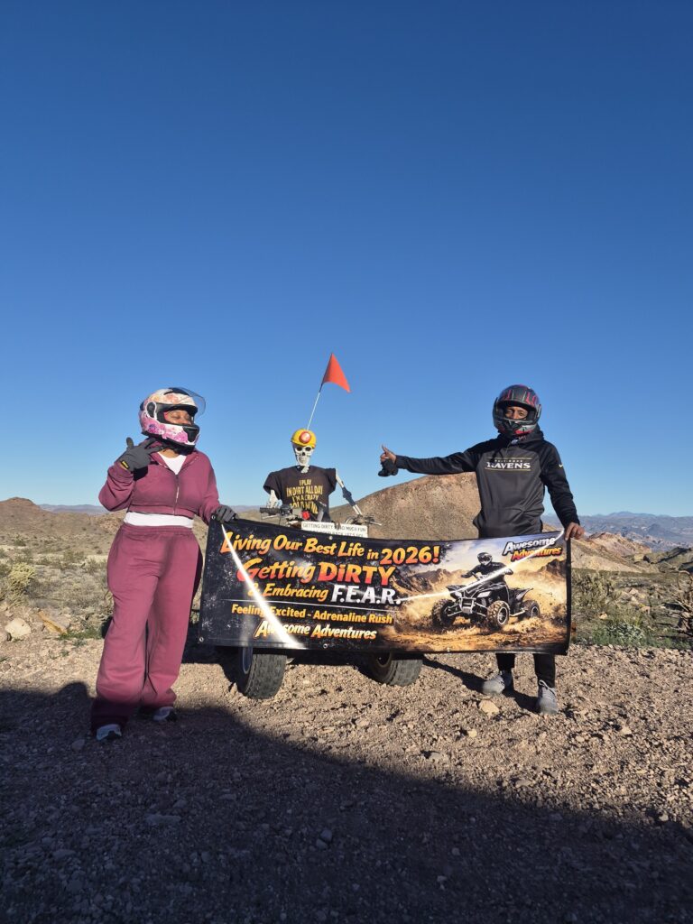 Three racers with helmets hold an off-road adventures banner near Nevada’s rocky terrain and mountains—perfect for Las Vegas ATV tours.