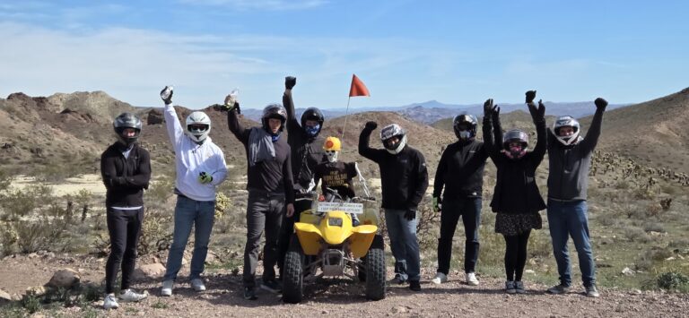 Group in helmets raises arms during a Las Vegas ATV tour in the Nevada desert; yellow RZR and hills in background.