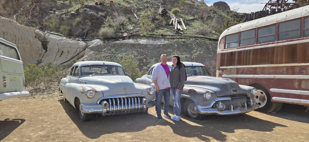 Smiling duo between vintage cars in Nevada desert near Las Vegas, with old bus and scenic hills—perfect for Ghost Town sightseeing.