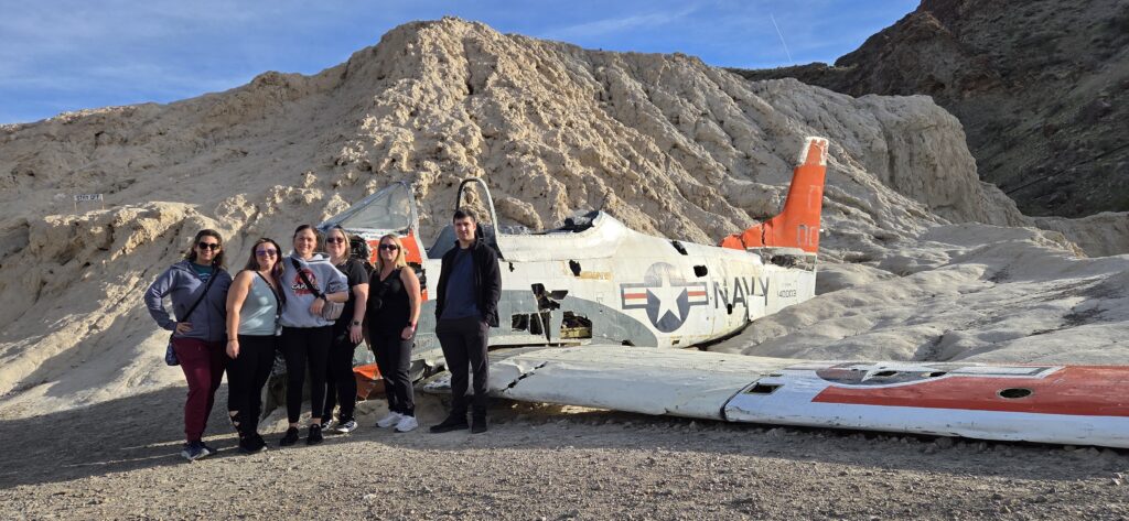 Group poses by a crashed US Navy plane in Nevada desert near rocky hills—perfect stop for Las Vegas ATV and Ghost Town tours.