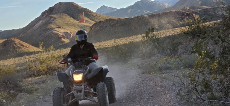 Rider in a helmet and black gear on an ATV explores a dusty Nevada desert trail near Las Vegas with mountains in the background.