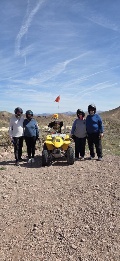 Group in helmets on a Nevada desert ATV tour near Las Vegas, parked by yellow ATV and orange flag under clear skies.