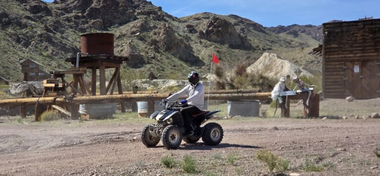 ATV rider explores a rustic Nevada trail near Las Vegas, passing wooden ghost town buildings and a red water tank under blue skies.