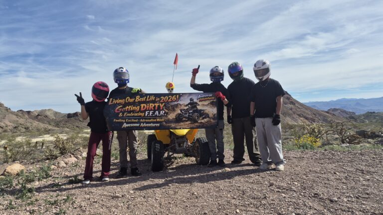 Group in helmets and off-road gear with ATV, holding an adventure banner near Las Vegas desert mountains on an ATV tour.