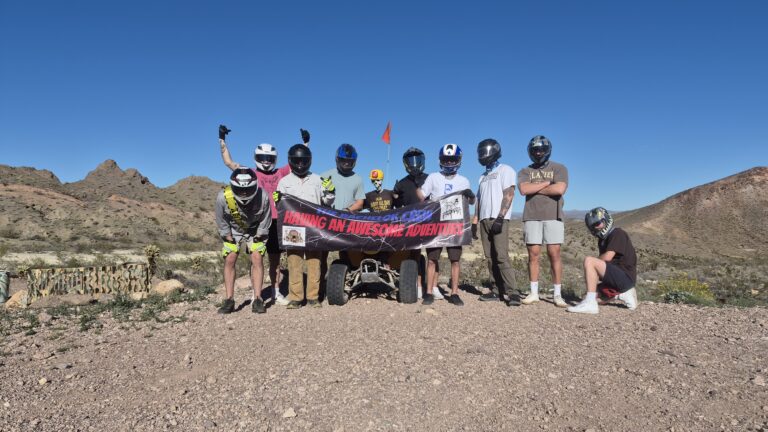 Group in helmets poses with WINNING AN AWESOME ADVENTURE banner on a Las Vegas ATV tour, desert hills and ATV in background.