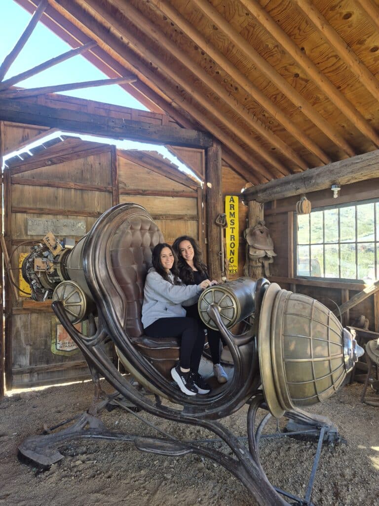 Two women smile on a steampunk chair in a rustic Nevada ghost town building, sunlight streaming in—perfect for sightseeing tours.