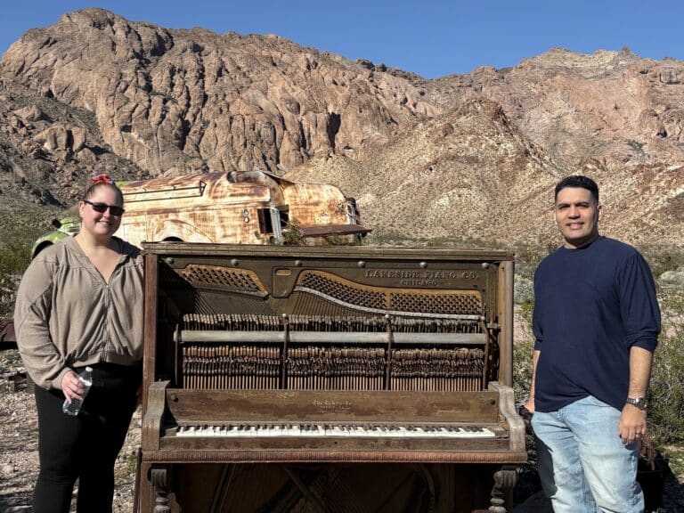 Two people by an old piano in Nevada’s desert near Las Vegas, with rocky hills, ghost town sights, and abandoned vehicles.