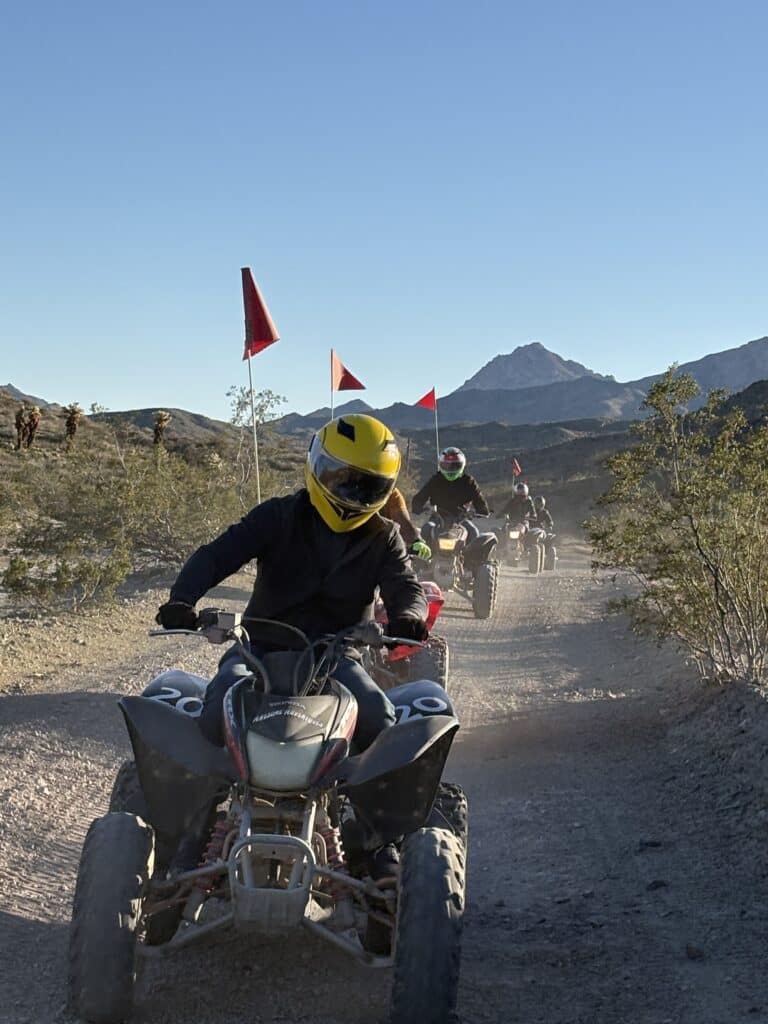 Group on ATV tour near Las Vegas rides a dusty desert trail with red flags, helmets, rocky Nevada hills, and sparse vegetation.