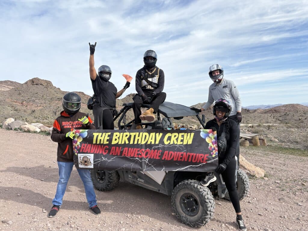 Group in helmets pose by an ATV in Nevada desert near Las Vegas, holding “Birthday Crew” banner on an off-road RZR adventure.