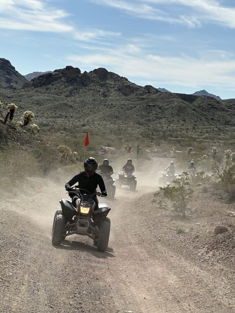 ATV tour group rides a dusty Nevada desert trail near Las Vegas, mountains visible, lead rider in front, under partly cloudy sky.