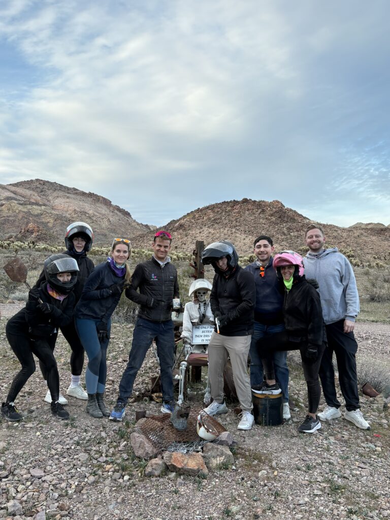 Group of 8 enjoy RZR off-road ride near Las Vegas, Nevada, posing by a skeleton in desert mountains under partly cloudy skies.