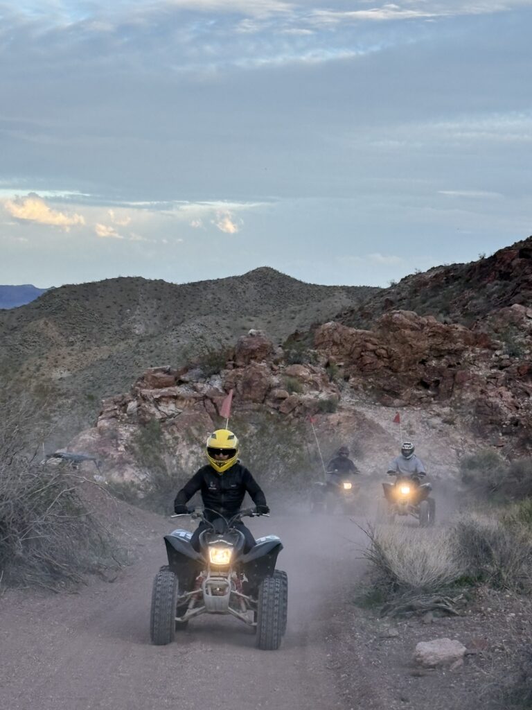 ATV tour riders in Nevada’s rocky desert near Las Vegas, with a yellow-helmet leader on a dusty trail under partly cloudy skies.