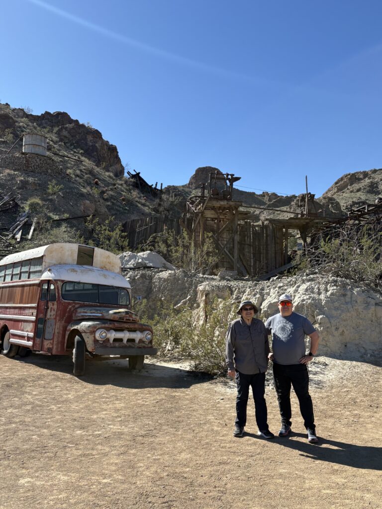 Two people explore a Nevada ghost town near Las Vegas, posing by a rusty bus and old wooden shacks—perfect for ATV tours.