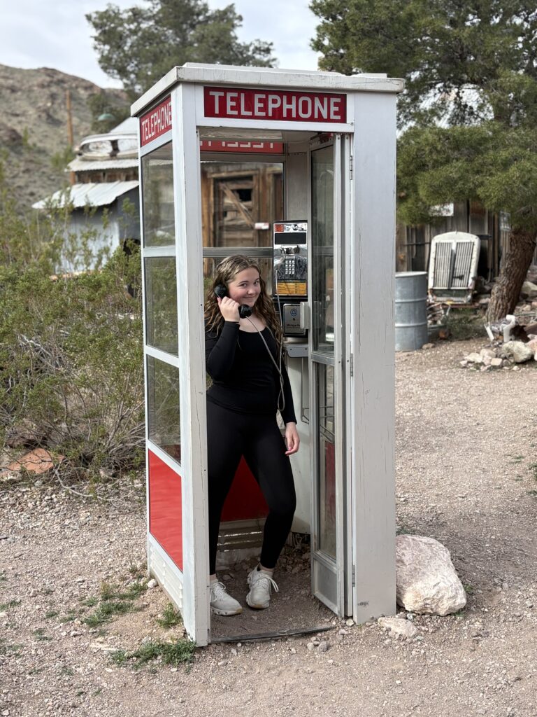 Smiling woman in a vintage phone booth at Nevada ghost town near Las Vegas, surrounded by trees and rustic buildings.